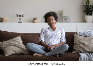 Smiling Millennial African American Woman Sit Rest On Couch In Living Room Watch TV On Weekend Break. Happy Young Ethnic Female Renter Tenant Relax On Sofa At Home Enjoy Television Program Online.