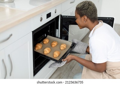 Smiling Millennial African American Woman Chef Taking Out Croissants From Oven In Kitchen Interior, Profile, Copy Space. Food Blog, New Recipe For Cooking And Baking At Home During Covid-19 Outbreak