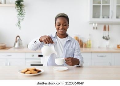 Smiling Millennial African American Woman In Casual Pouring Milk At Coffee At Table Minimalist Kitchen Interior. Food Blog, Housewife Rest At Home, Visit And Healthy Breakfast During Covid-19 Outbreak