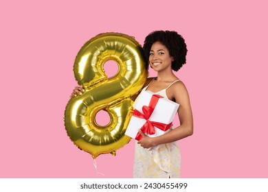 Smiling millennial african american woman with a natural afro hairstyle holding a golden number 8 balloon and a white gift box with a red ribbon against a pink backdrop, studio