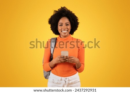 Smiling millennial African American woman with curly hair, wearing an orange turtleneck, texts on her phone while carrying a grey backpack on a vibrant yellow background, studio