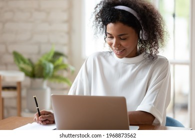 Smiling millennial african ethnicity woman in headphones listening educational online lecture, writing notes or holding web camera video call distant negotiations with partners in modern office.
