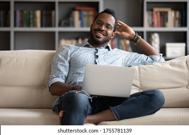 Smiling millennial African guy sit on couch in living room working on wireless pc from home, happy young man resting on sofa browsing internet shopping using computer, modern technology usage concept - Shutterstock ID 1755781139