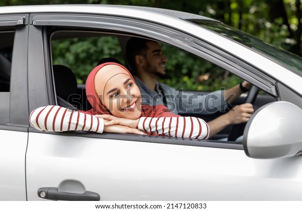 Smiling millennial arab female in hijab ride in\
car looks out the open window outdoor, enjoy journey, man drives\
car. Travel in summer, weekends on road, holidays with family after\
covid-19 pandemic