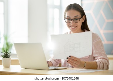 Smiling millennial architect working with architectural house plan drawing and laptop, creative young female professional interior designer holding new project blueprint planning design for client