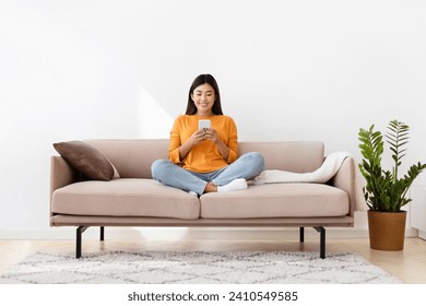 Smiling millennial asian lady holding smartphone, sitting on couch in cozy living room, home interior. Filipino woman reading online book or blog on cell phone, copy space, full length - Powered by Shutterstock