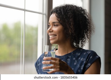 Smiling Millennial Biracial Woman Look In Distance Hold Glass Of Water Drink Clean Clear Mineral Aqua For Body Refreshment. Happy African American Girl Follow Healthy Lifestyle. Hydration Concept.