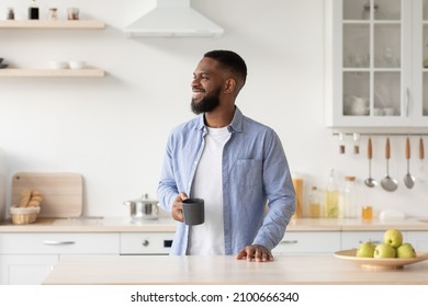 Smiling millennial black male with beard enjoys good morning and fresh coffee in cup and looks at free space at minimalist kitchen interior. Relax, break, breakfast with favorite drink, ad and offer - Shutterstock ID 2100666340