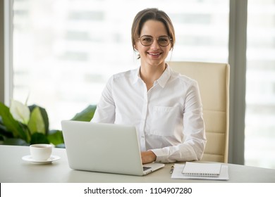 Smiling millennial businesswoman in glasses sitting at work desk with laptop, friendly professional looking at camera at workplace, female boss, company executive or successful entrepreneur portrait