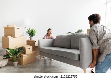 Smiling Millennial Caucasian Couple In Casual Carry Sofa In Room With Packed Cardboard Boxes, Planning Interior. Buy New Furniture, Moving Together At Renting House, Own Apartment And Family Property