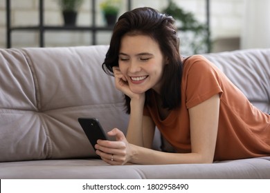 Lächelnde Jahrtausende kaukasische Weibchen entspannen sich auf Sofa Text-Nachricht auf dem Handy. Fröhliche junge Frau, die sich auf einem Sofa im Wohnzimmer zu Hause ruht, surfen im drahtlosen Internet auf dem Smartphone. Technologiekonzept