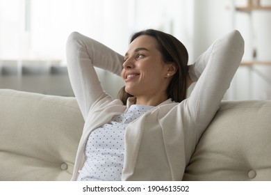 Smiling millennial Caucasian female relax take break on comfortable sofa at home relieve negative emotions dreaming. Happy young woman renter or tenant rest on couch breathe fresh air. Peace concept.