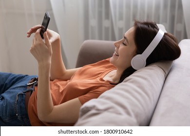 Smiling millennial Caucasian girl in headphones relax on couch at home play use modern smartphone gadget, happy young woman in earphones rest on sofa in living room browse internet text on cellphone - Powered by Shutterstock