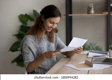 Smiling Millennial Caucasian Girl Sit At Table At Home Read Good News In Postal Letter Correspondence, Happy Young Woman Open Envelope Feel Excited Get Pleasant Message Or Notice In Post Paperwork