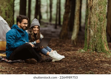 Smiling millennial caucasian guy and woman hikers in jackets relaxing in cold forest, enjoy adventure, sit in tent, typing on laptop outdoor. Travel blog, work, vacation, active lifestyle and camping - Shutterstock ID 2264989753