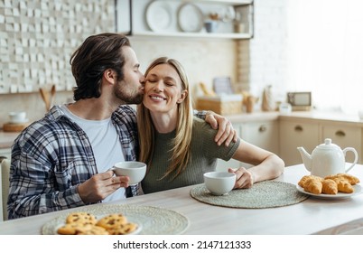 Smiling Millennial Caucasian Husband With Stubble Hugs And Kisses His Wife On Cheek, Couple Drinks Coffee In Modern Kitchen Interior. Free Time, Breakfast With Drink Together And Good Morning At Home