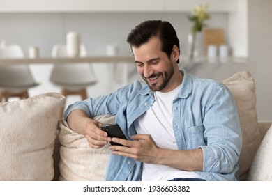 Smiling Millennial Caucasian Man Sit On Couch At Home Have Fun Using Modern Cellphone Gadget. Happy Young Male Relax On Sofa Look At Smartphone Screen Texting Or Messaging Online. Technology Concept.