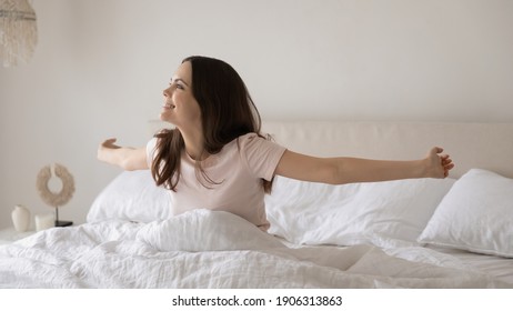 Smiling millennial Caucasian woman wake up sit in comfortable bed meet new sunny day. Happy young female stretch awaken in cozy bedroom at home, enjoy early weekend morning after good sleep.