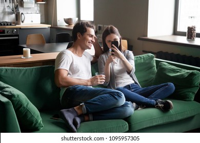 Smiling Millennial Couple Enjoying Watching Mobile Video On Phone Sitting On Sofa At Home, Positive Teenage Girl Holding Smartphone Showing Funny Online Photo To Boyfriend Spending Weekend Together