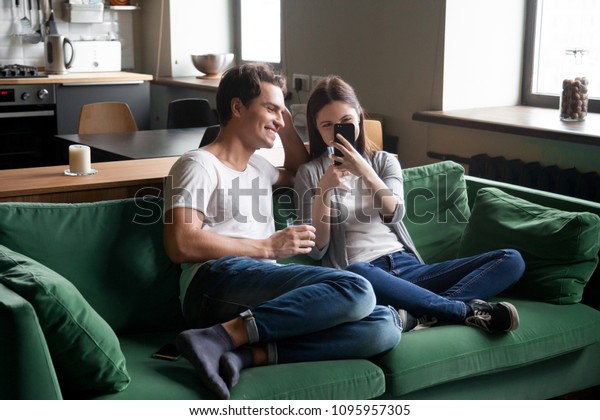 自宅のソファ に座って携帯電話でビデオを見るのを楽しむ00年生の笑顔のカップル 楽しいオンライン写真を持つ10代のポジティブな女の子が週末を一緒に過ごす彼氏のために 楽しいオンライン写真を持つ の写真素材 今すぐ編集