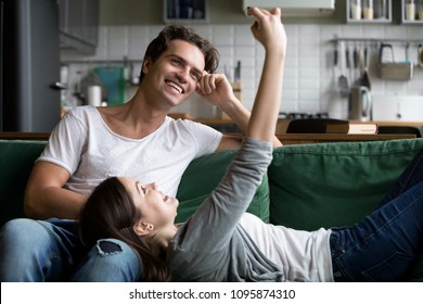 Smiling Millennial Couple Having Fun With Smartphone Taking Selfie At Home, Happy Teen Girl Relaxing On Couch With Boyfriend Holding Cellphone Using Mobile Apps At Home Making Photo Or Shooting Blog