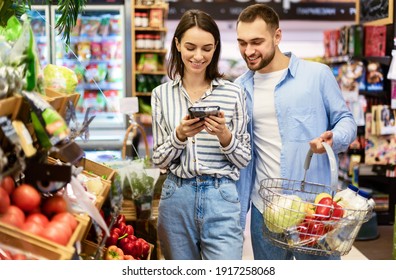 Smiling Millennial Couple Shopping Groceries In The Hypermarket, Two Happy Consumers Buying Food Walking With Cart In Wholesale Shop, Choosing Eco Vegetables And Fruits, Checking Label And Expiry Date