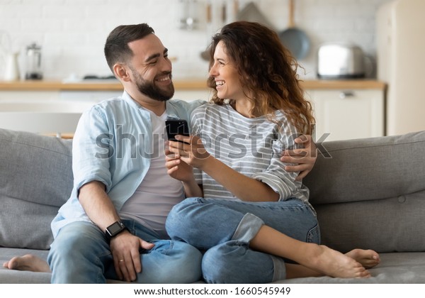 Smiling Millennial Couple Sit On Couch In Kitchen Have Fun Browsing Internet On Modern
