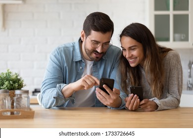 Smiling millennial couple sit at table in kitchen have fun using modern smartphone devices together, happy young husband and wife laugh relax at home browsing application on cellphone gadgets