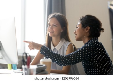 Smiling millennial diverse colleagues discussing online work together, happy caucasian female intern listening to indian mentor explaining computer task training employee, office mentoring concept