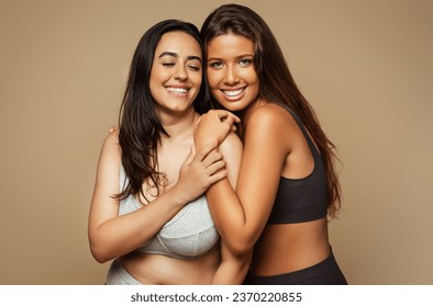Smiling millennial diverse women hugs in underwear enjoy health, beauty care isolated on beige background, studio. Love for self, motivation, spa treatments wellness, skin care of different types Stock fotografie