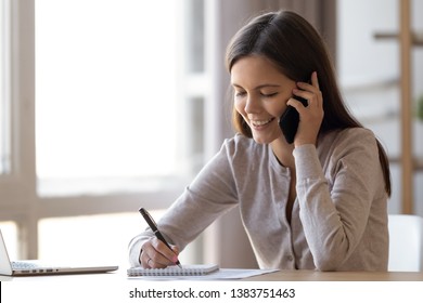 Smiling millennial girl distracted from studying at laptop talk chat over phone drawing in notebook, happy young woman sit at office table having pleasant cellphone conversation, female flirt on cell
