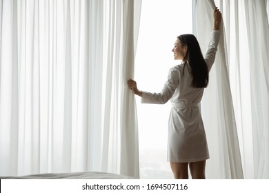 Smiling Millennial Girl Wearing Bathrobe Open Curtains In Bedroom Welcome New Sunny Day, Happy Positive Young Woman Wake Up From Sleep Look In Window At Home Or Hotel Feeling Optimistic