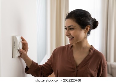 Smiling millennial indian ethnicity woman setting comfortable domestic temperature using modern technology controller. Happy female homeowner using smart home system, activating alarm.