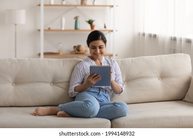 Smiling millennial Indian female renter relax on sofa in living room using modern tablet gadget. Happy young mixed race woman rest on couch at home shopping online on pad device. Technology concept.