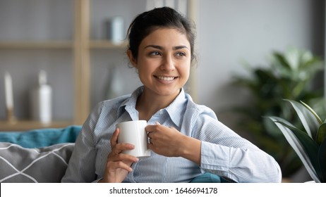 Smiling Millennial Indian Girl Relax On Couch In Living Room Drink Tea Dreaming Or Visualizing, Happy Dreamy Young Ethnic Woman Rest On Sofa Enjoy Warm Coffee Look In Distance Thinking At Home