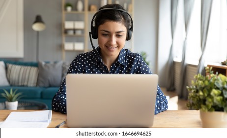Smiling millennial Indian girl in wireless headset sit at desk in living room study online on laptop, happy young ethnic female in headphones watch webinar or course on Internet on computer