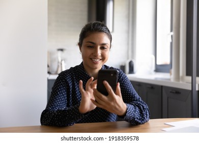 Smiling Millennial Indian Woman Sit At Desk At Home Look At Smartphone Screen Texting Messaging Online. Happy Young Ethnic Female Use Cellphone Browse Wireless Internet On Gadget. Technology Concept.