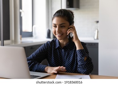 Smiling millennial Indian woman work on laptop from home office talk on cellphone consulting client or customer online. Happy young ethnic businesswoman have smartphone consultation with partner.