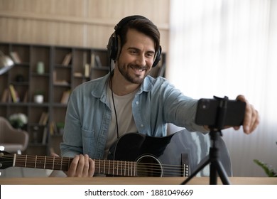 Smiling millennial male artist blogger playing on guitar shooting music video or tutorial on modern smartphone at home studio. Happy young man singer use music instrument, compose song on cellphone. - Powered by Shutterstock