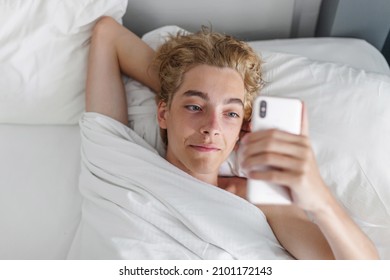 Smiling Millennial  Male Awake Lying In White Bed Checking Mail On Smartphone, Happy Young Man Wake Up In Morning Using Cellphone Get Pleasant Message Or Text. Smiling Teen On Bedroom With Phone