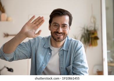 Smiling millennial male in glasses posing before digital webcam waving hand chatting with friend using pc. Headshot portrait of active young man video blogger looking at camera broadcasting from home - Shutterstock ID 1910861455