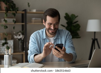 Smiling millennial man sit at table at home office look at smartphone screen texting or messaging. Happy young Caucasian male browse surf wireless internet on cellphone. Communication concept.