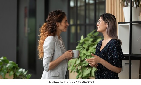 Smiling millennial multiracial female employees talk chat during coffee break in office, young multiethnic women colleagues speak laughing enjoy leisure time at workplace, cooperating concept