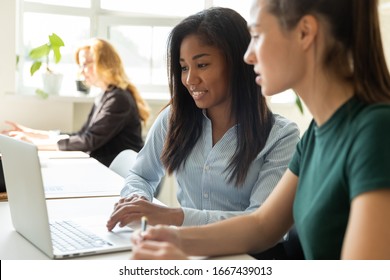 Smiling millennial multiracial female employees cooperate working together on laptop in shared office, diverse women coworkers busy collaborating discuss ideas brainstorm at business meeting