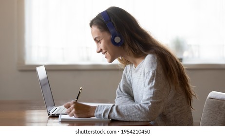 Smiling millennial teen female in headphones look at laptop screen write study online at home. Happy young Latin woman in earphones handwrite make note work distant on computer. Education concept.