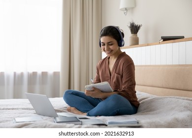 Smiling millennial teen Indian female student in headphones sit on bed at home take note study online on computer. Happy young ethnic woman in earphones busy take distant course or training on laptop.