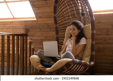 Smiling Millennial Woman In Pajamas Relax In Cozy Chair At Home Look At Laptop Screen Talk On Video Call. Happy Young Female Rest In Comfy Country House Watch Movie On Computer On Leisure Weekend.