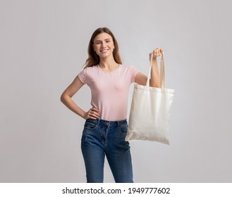 Smiling Millennial Woman Posing With Blank Eco Tote Canvas Bag, Demonstating Free Copy Space For Your Design Or Logo, Positive Young Lady Standing Over Light Studio Background, Looking At Camera