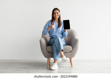 Smiling millennial woman showing empty touch pad, gesturing thumb up, sitting in armchair against white wall, mockup for website design on tablet screen. Online ad concept - Shutterstock ID 2021599880