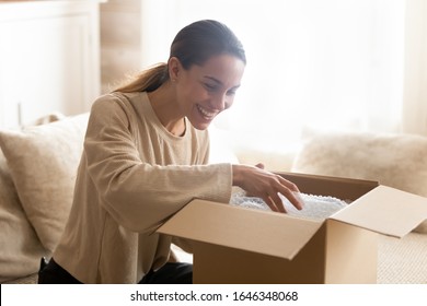 Smiling Millennial Woman Sit On Couch At Home Open Post Package Shopping Online Buying Goods On Internet, Happy Young Female Customer Unpack Postal Shipping Parcel Satisfied With Order Or Delivery